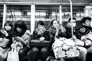 A group of people sitting on a subway, all on their cell phones.