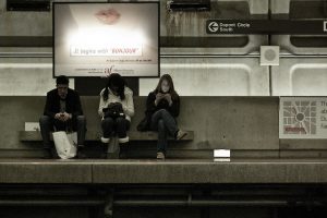 Three people sitting under a advert looking at their phone while waiting for a train.